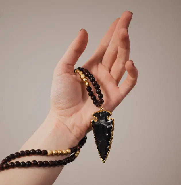 A hand elegantly presents a striking arrowhead-shaped pendant with a glossy black finish, edged in gold, suspended from a beaded necklace featuring hues of deep brown and subtle gold, exemplifying bespoke craftsmanship.