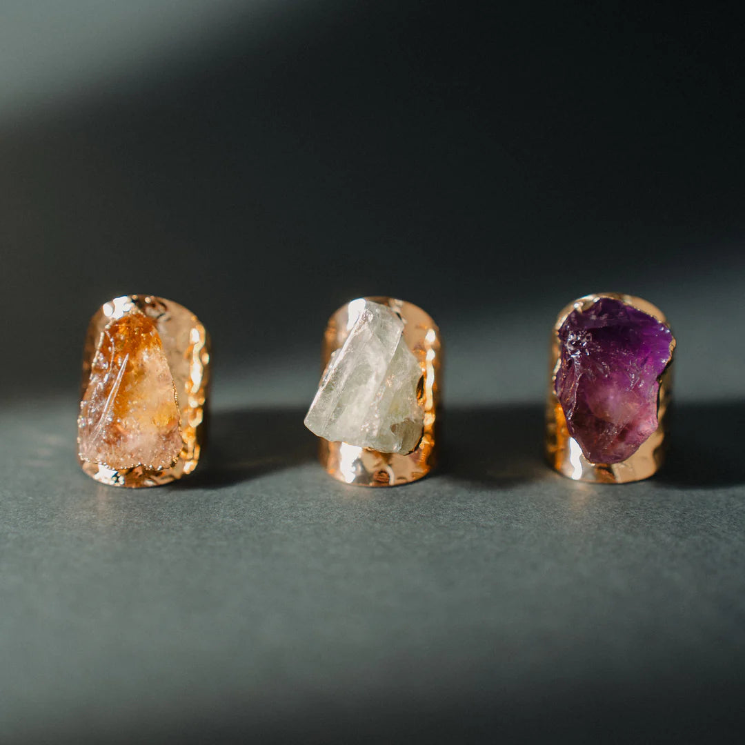 Three raw crystal rings are displayed against a dark backdrop, featuring natural, uncut stones in warm amber, clear quartz, and deep purple amethyst, each set in a gleaming copper band, highlighting the organic beauty of these healing crystal gems.